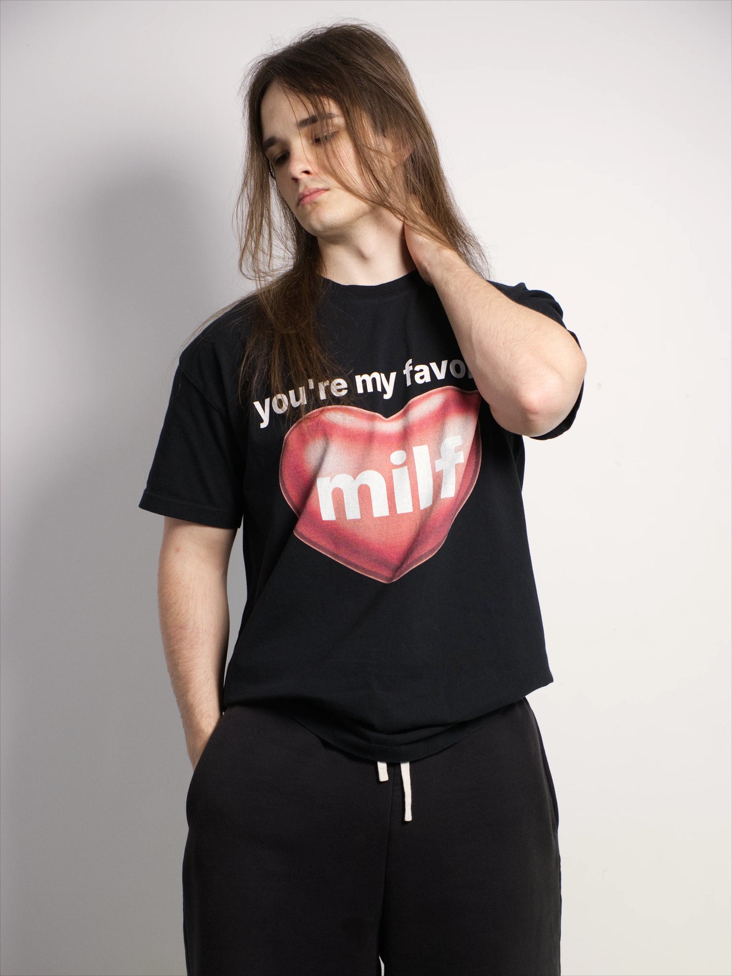 A man wearing a black graphic t-shirt with a red heart and white text that reads 'you're my fav milf', paired with black sweatpants, against a plain light gray background.