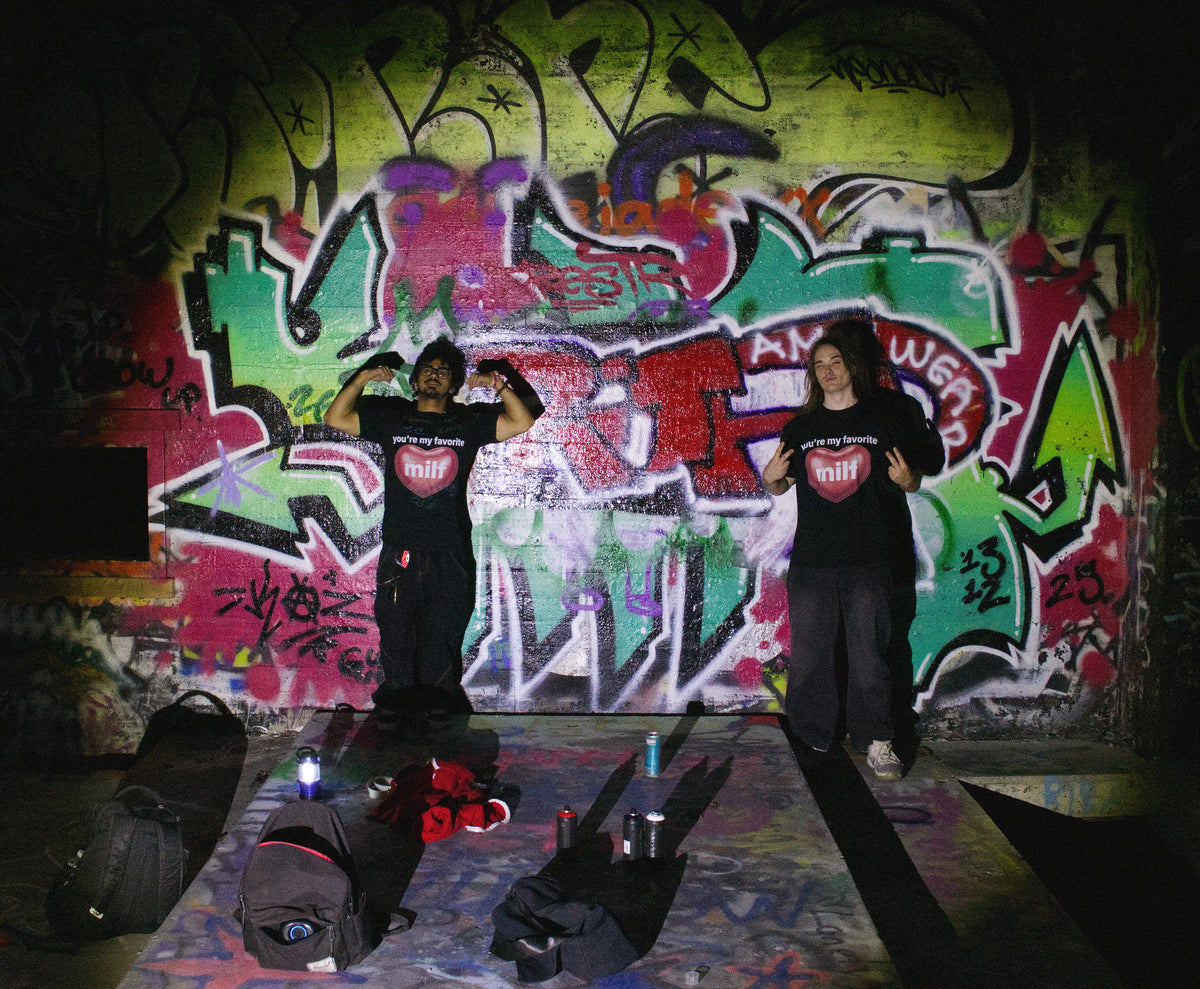 Two people posing in front of a vibrant graffiti wall, wearing matching black t-shirts with a red heart graphic and black sweatpants. Their bags and shoes are scattered on the floor in the foreground.